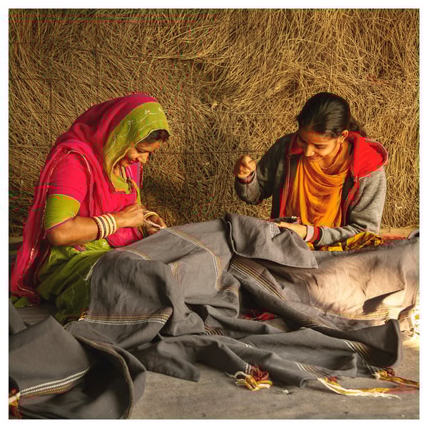 Two women in bright saris weaving intricate patterns on fabric outdoors.