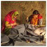 Two women in bright saris weaving intricate patterns on fabric outdoors.