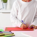 A child in a white sweater is drawing with a blue pen on red paper. Several colorful pens are nearby.