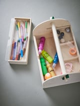 Two wooden storage boxes against a wall. One contains colorful pens. The other has art supplies, including paints, brushes, and tape.