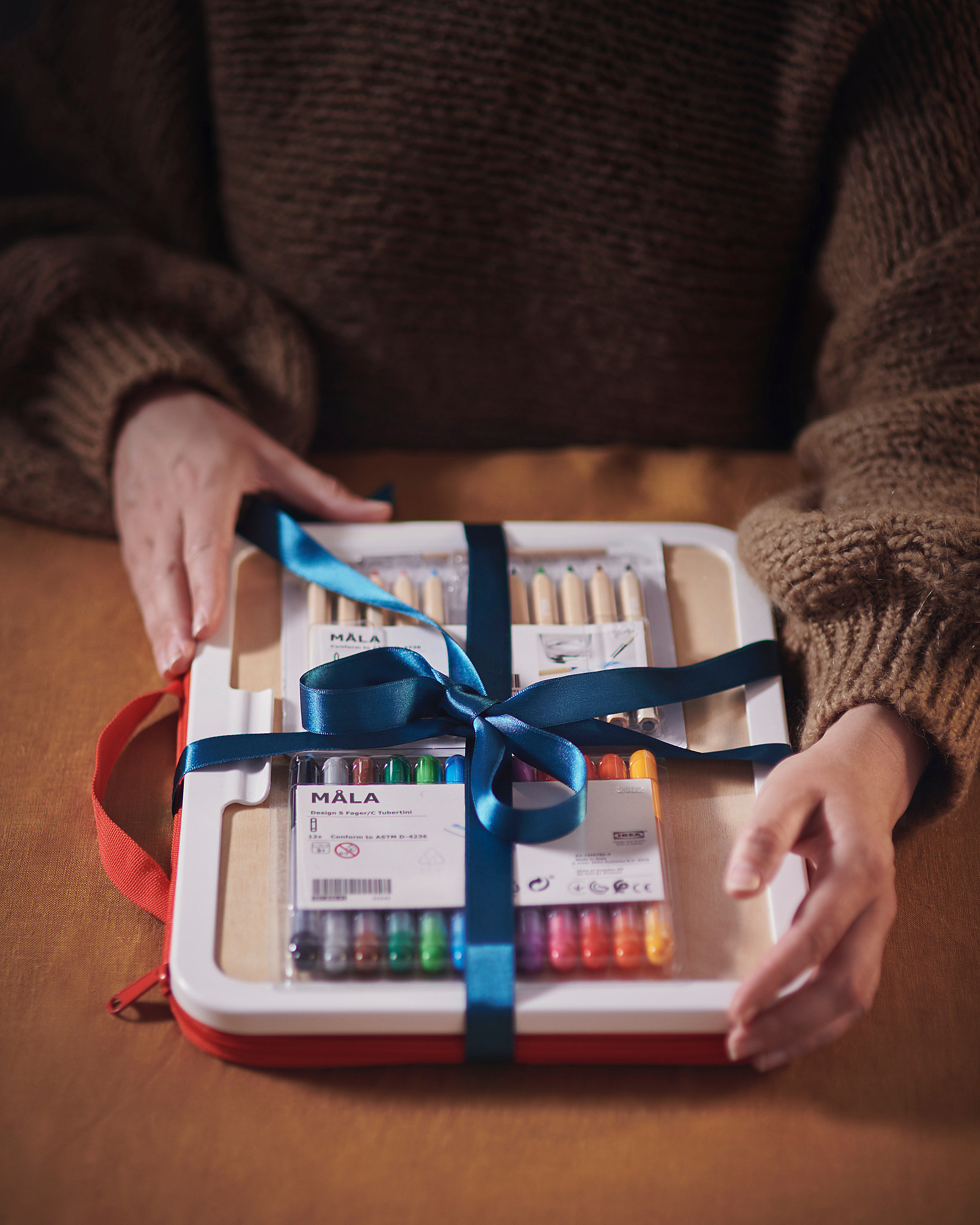 A person holding a MÅLA art set, white with a blue ribbon, containing markers and pens.