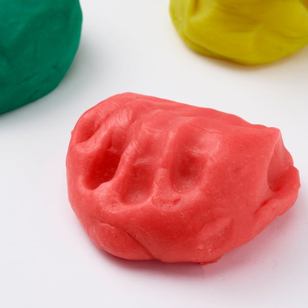A close-up of bright red play-doh, with green and yellow clumps in the background, showcasing its smooth, pliable texture.
