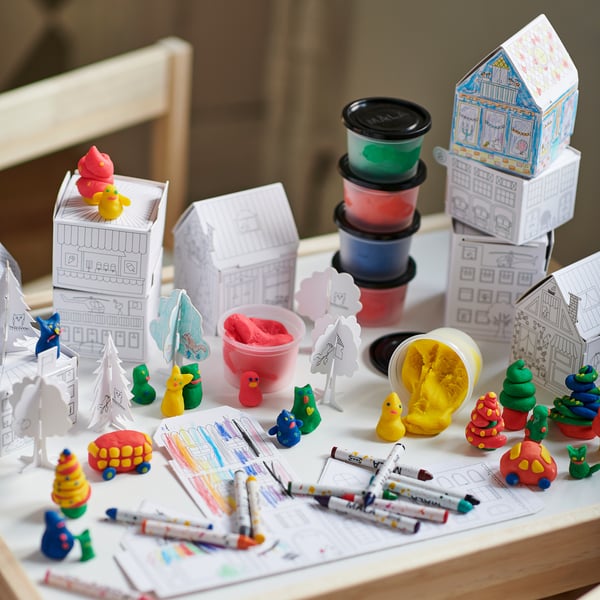 A childs craft table with MÅLA colorful modeling dough, pens, paper houses, and small toys in various shapes and colors.