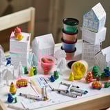 A childs craft table with MÅLA colorful modeling dough, pens, paper houses, and small toys in various shapes and colors.
