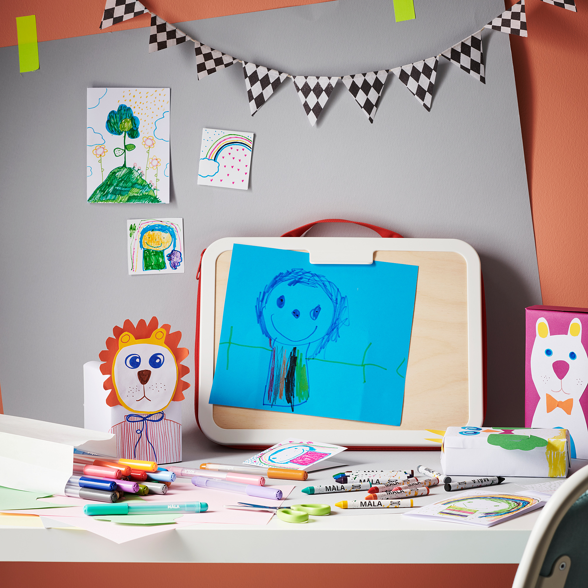 Art station with MÅLA white box holding blue paper featuring a drawing of a girl. Surrounded by colorful pens, markers, and paper crafts on a desk.