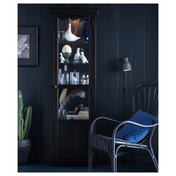 Black glass cabinet with decor and blue cushion in wicker chair beside cactus.