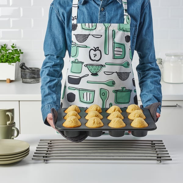 Person holding a muffin tin with baked muffins, wearing a denim shirt and an apron with kitchen tools pattern.