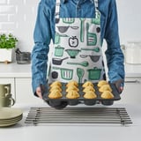 Person holding a muffin tin with baked muffins, wearing a denim shirt and an apron with kitchen tools pattern.