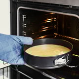 A hand in a blue oven mitt places a round cake pan with a golden batter into an open oven, showcasing its non-stick and easy-release.