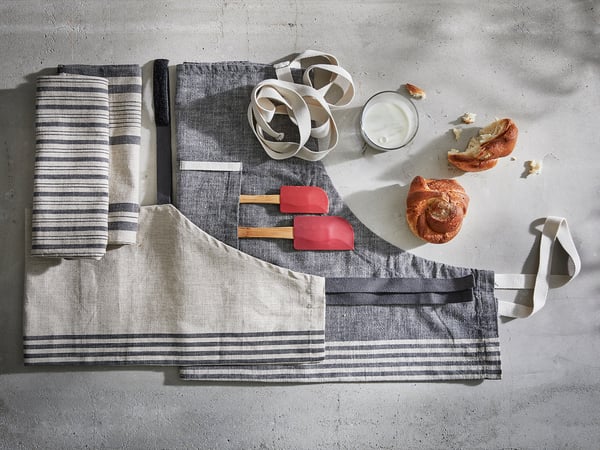 A gray linen-cotton apron with stripes and pockets rests on a gray concrete surface, accompanied by kitchen tools and pastries.