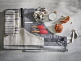 A gray linen-cotton apron with stripes and pockets rests on a gray concrete surface, accompanied by kitchen tools and pastries.