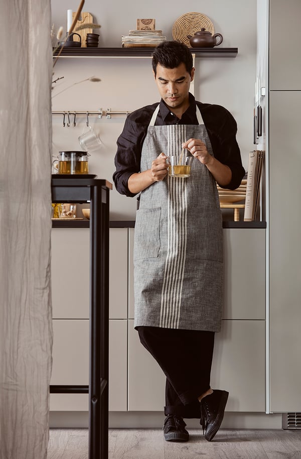 Man wearing black shirt and gray apron holds cup in modern kitchen. Apron has stripes and pockets. Shelves with items behind.