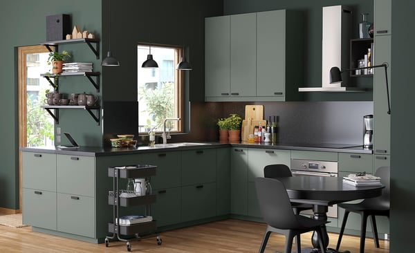 Modern kitchen with greenish-gray cabinets, black appliances, and wood floor. Shelves, potted plants, and black chairs around a small table.