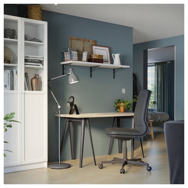 A grey ikea MITTCIRKEL desk setup in a blue room. It a light pine-like top and adjustable chair, with a bookshelf and lamp nearby.