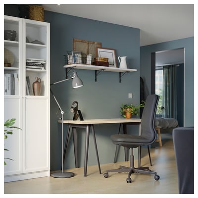 A grey ikea MITTCIRKEL desk setup in a blue room. It a light pine-like top and adjustable chair, with a bookshelf and lamp nearby.