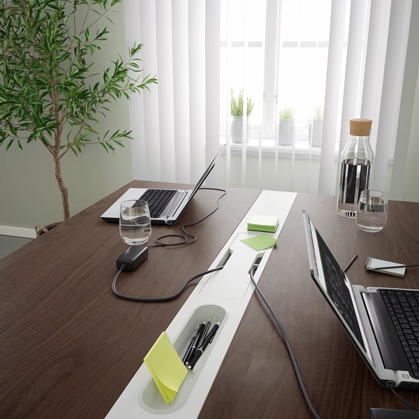 Modern conference table with laptops, glassware, and plants. Cable access and storage compartments. Bright and airy setting with large windows.