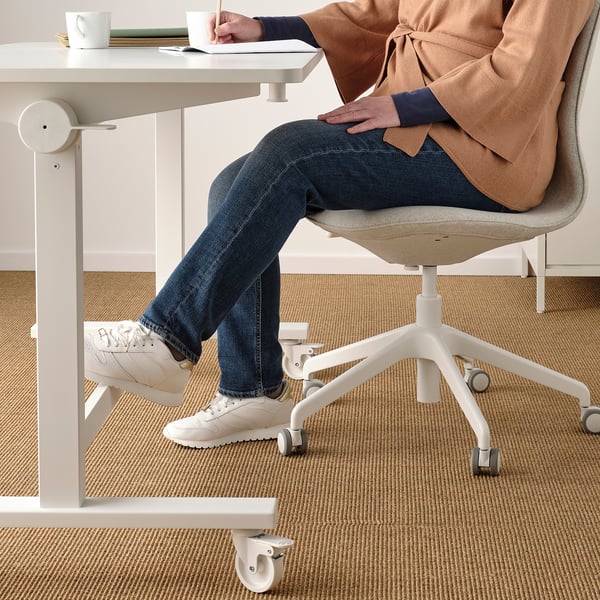 Person sitting at adjustable table with footrest, taking notes. White sneakers, jeans, and sweater visible.