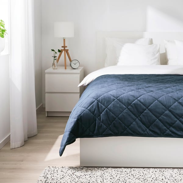 Minimalist bedroom with white walls, wooden floor, blue velvet bed, bedside lamp, and clock.