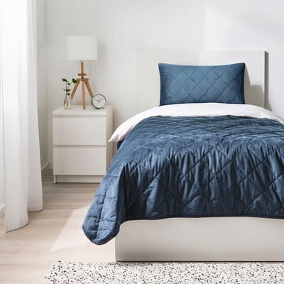 Tiny bedroom: white bed with dark blue velvet cushion, wooden nightstand with lamp & clock.
