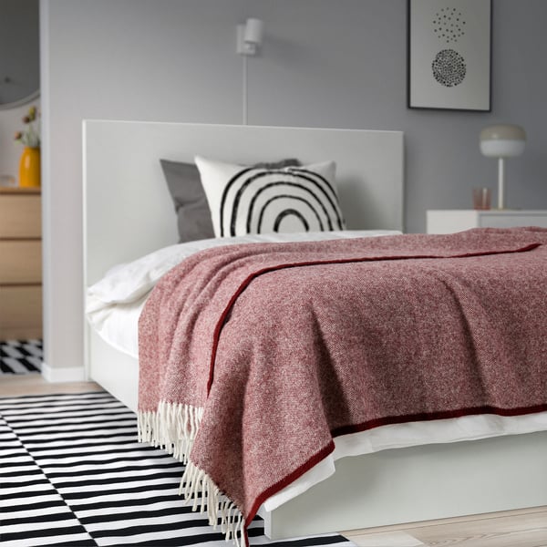 A maroon wool throw blanket with fringes on a bed. White headboard, black & white rug. Geometric pillow, grey wall with art. Side table with lamp.