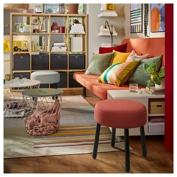 Bright living room with orange sofa, colorful pillows, striped rug, wire furniture, and plants. Centered orange stool.