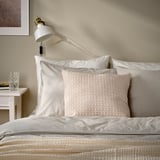 A MOROTSMOTT pillow on a bed. The pillow is light pink with a woven pattern. Its smaller than the bed and surrounded by larger white pillows.