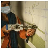 Person tufting beige-green rug with white gun, orange sweater, blue mask.