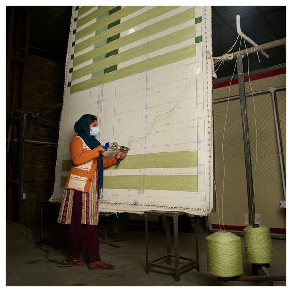 Woman crafting large striped rug, using green, white, black yarn on loom, wearing orange, black clothing, in industrial setting.