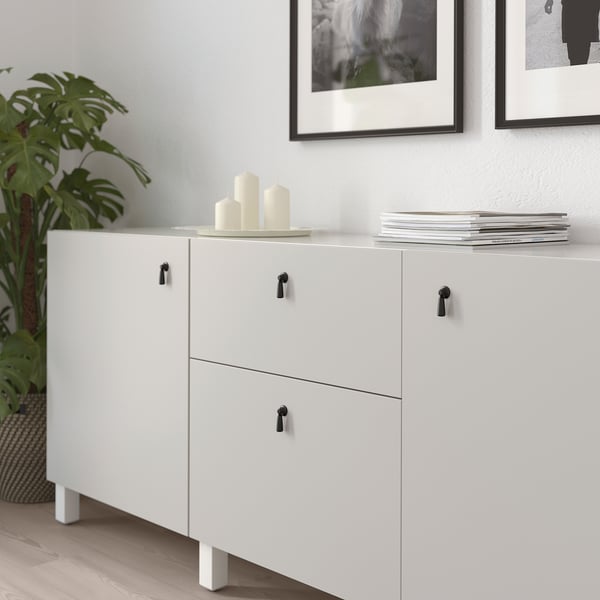 White cabinet with black drop handles; three drawers, two doors, decorative candles and books on top.