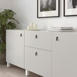 White cabinet with black drop handles; three drawers, two doors, decorative candles and books on top.