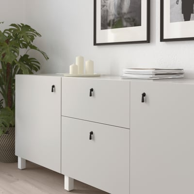 White cabinet with black drop handles; three drawers, two doors, decorative candles and books on top.