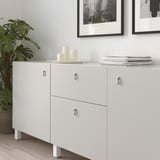 White cabinet with drop handles preventing damage, three candles, stack of magazines, framed pictures, green plant.
