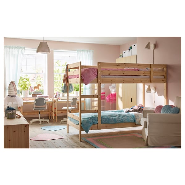 Shared kids room with pink walls, wooden bunk beds, desks, and storage shelves. Bed ceiling light bulb, toys, and pastel bedding.