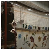 Man weaving vibrant, flower-patterned rug on loom.