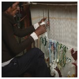 Man weaving rug. Brown sweater, jeans. Threads around loom.