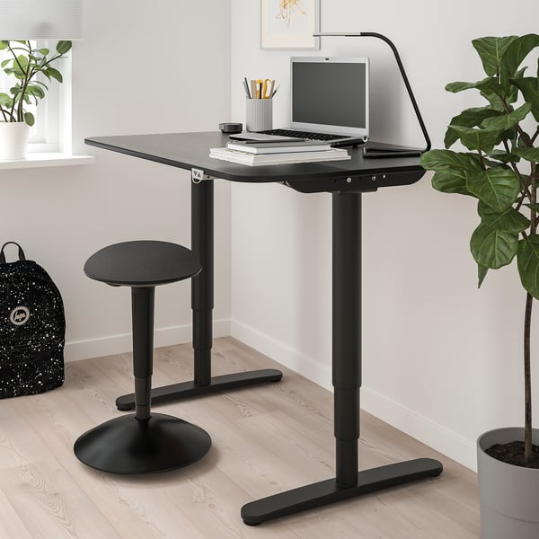 Modern black home office setup with height-adjustable desk & stool, laptop, books, lamp & green plants.