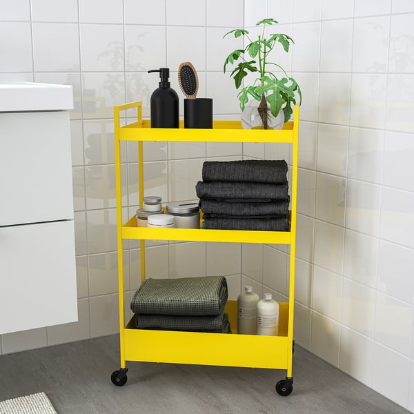 Yellow NISSAFORS storage trolley with black accents, holds towels, bottles, and a plant, fits in small spaces.