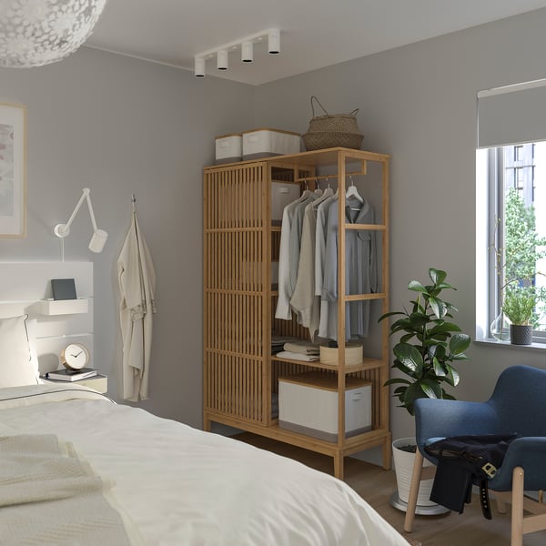 Minimalist grey bedroom with wooden wardrobe, white bed, and blue chair. Clothes and plants decorate the room.
