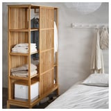 A NORDKISA wooden wardrobe with shelves filled with towels and clothing, placed beside a bed with white bedding.