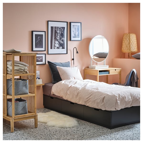 A cozy bedroom with a NORDLI bed in light wood and black storage drawers, set against a peach wall, with a white mirror and a wooden shelf beside it.