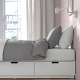 A cozy bed with grey linens sits by a window, offering both comfort and storage with its built-in drawers. The room soft lighting and sheer curtains.