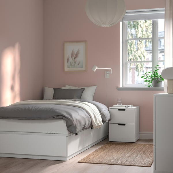 Minimalist bedroom with white bed, grey linens, pink walls, and plant.