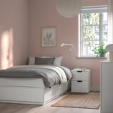 Minimalist bedroom with white bed, grey linens, pink walls, and plant.