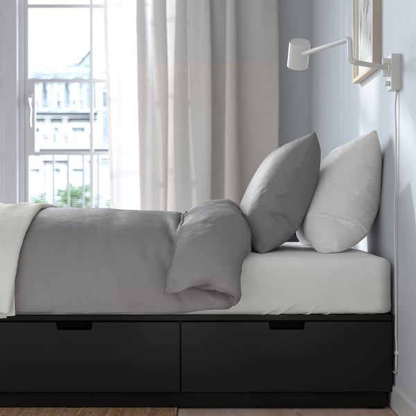 Modern bed with drawers under frame, grey pillows, light grey curtains, cityscape view from window.