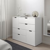 White NORDLI 6-drawer chest in bedroom between window and bed, modern minimalist style.
