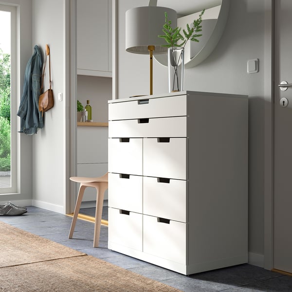 Modern white NORDLI chest with six drawers, tall, sleek, by door, near coat hooks and stool.