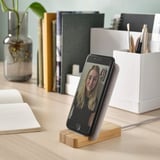 Wireless charging stand in white with bamboo base, smartphone on desk with books and plants.