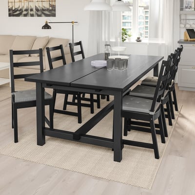 Black NORDVIKEN dining set with extendable table and 6 chairs. Modern-rustic, seats 8. Cozy, light-filled room with couch and lamp.