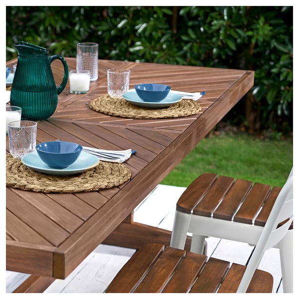 Outdoor dining setup with acacia table, stacked chairs, plates, bowls, glasses, and a green pitcher on placemats.
