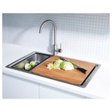 Modern stainless steel kitchen sink with wooden cutting board, cabbage, knife, and metal faucet against white tiled wall.